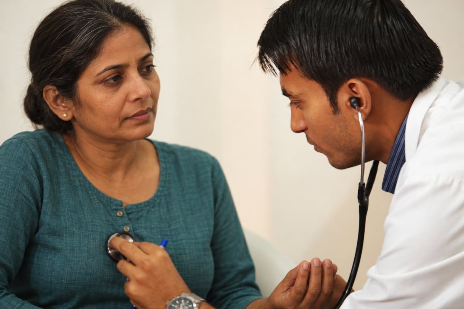 A doctor listening to a woman's heart with a stethoscope as she sits in a teal kurta