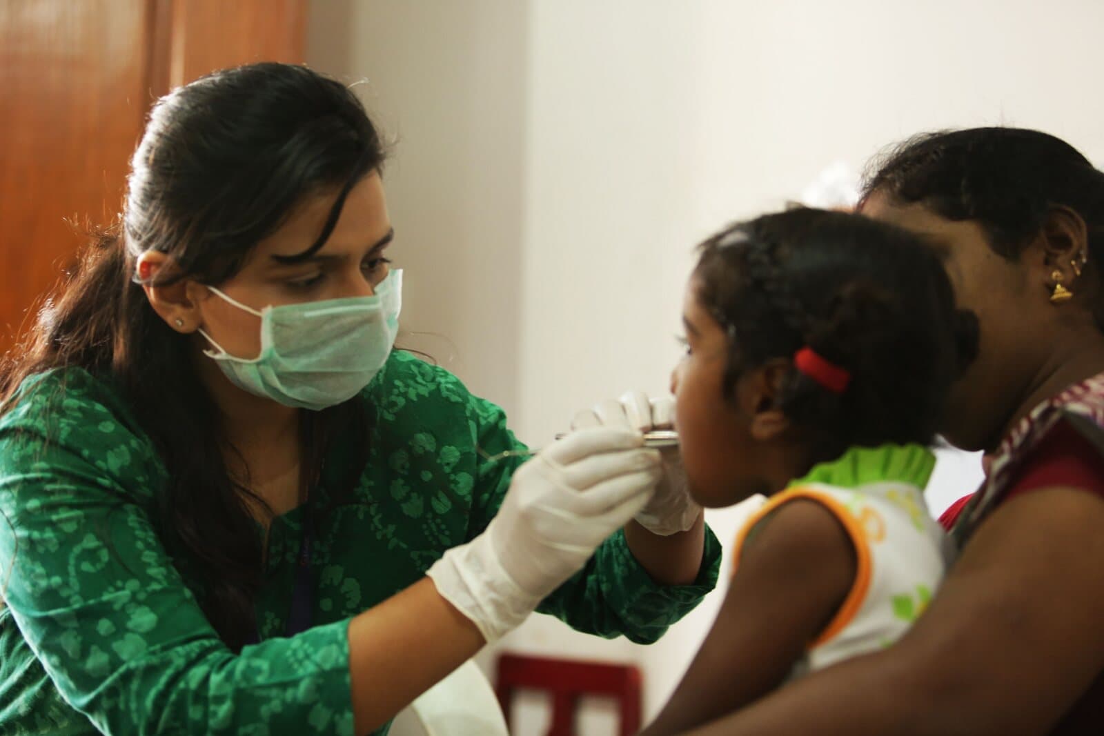 A doctor in a mask checking a young girl's mouth, her mother beside her